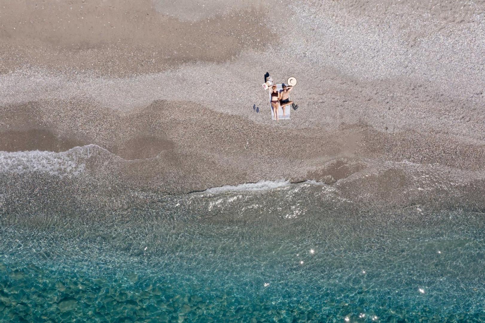 Aerofoto skats uz Alianthos Garden Hotel piekrasti — divi atpūtnieki pludmalē pie kristāldzidra, tirkīzzila ūdens Krētā
