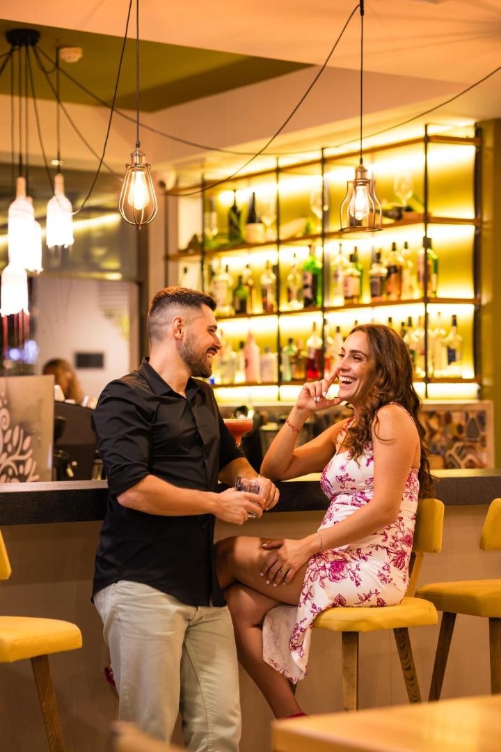 Pāris bauda dzērienus Alianthos Garden Hotel iekštelpu bārā — mājīgs apgaismojums, dzelteni bāra krēsli un plaša dzērienu izvēle fonā