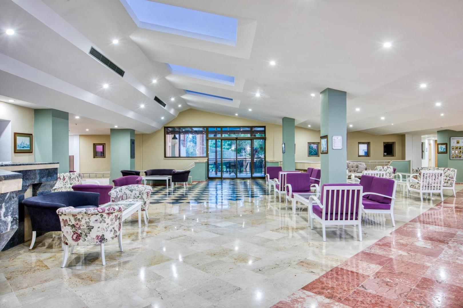 Asteria Collection Side viesnīcas lobby zona ar gaišām flīžu grīdām, violetiem atpūtas krēsliem un stikla jumta logiem