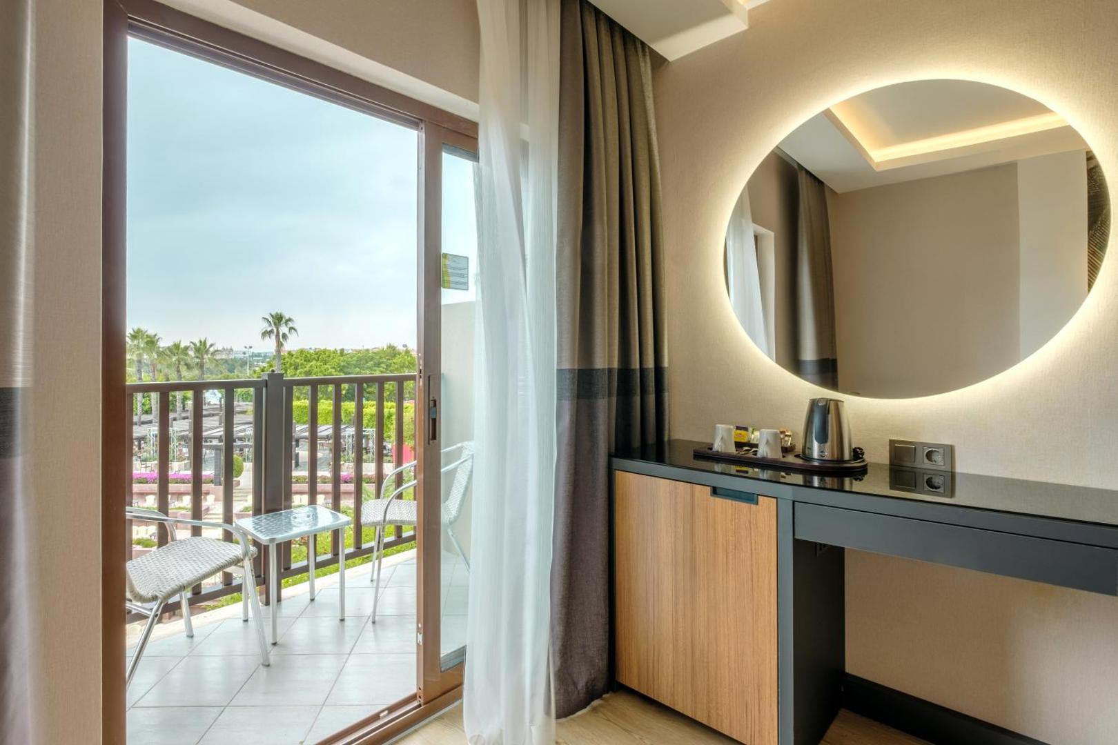 Alba Resort Hotel numura balkons ar skatu uz dārzu un palmām, apaļš izgaismots spogulis un kafijas pagatavošanas stūrītis