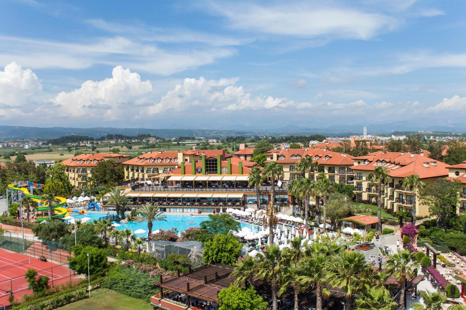 Alba Resort Hotel panorāmas skats uz viesnīcas korpusiem, baseinu un palmu dārzu uz kalnu fona Sidē.