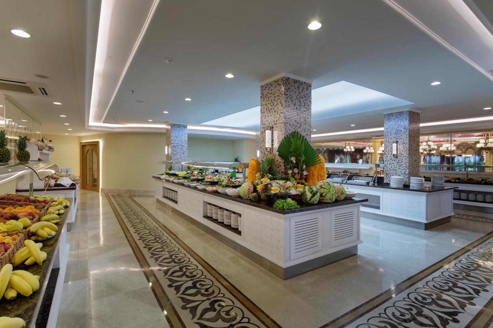 Alba Resort Hotel galvenā restorāna bufetes zona ar plašu augļu izvēli un dekoratīviem grebumiem gaišā, modernā interjerā