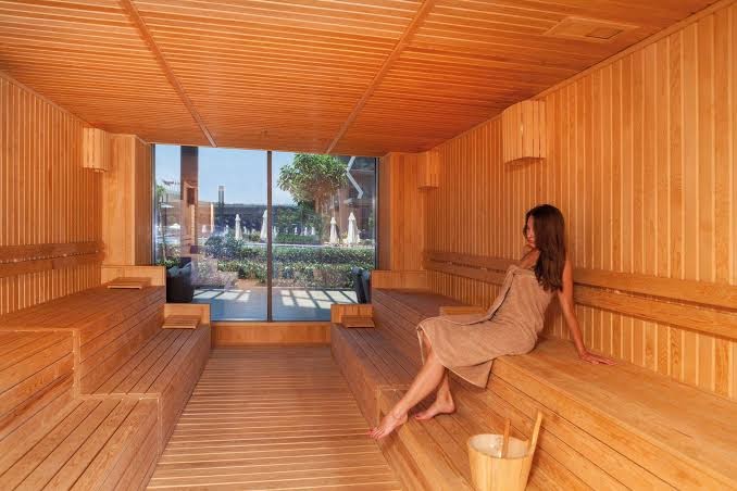 Bosphorus Sorgun Hotel spa zonas sauna ar koka apdari un lielu logu uz dārzu — sieviete atpūšas uz koka lāvām