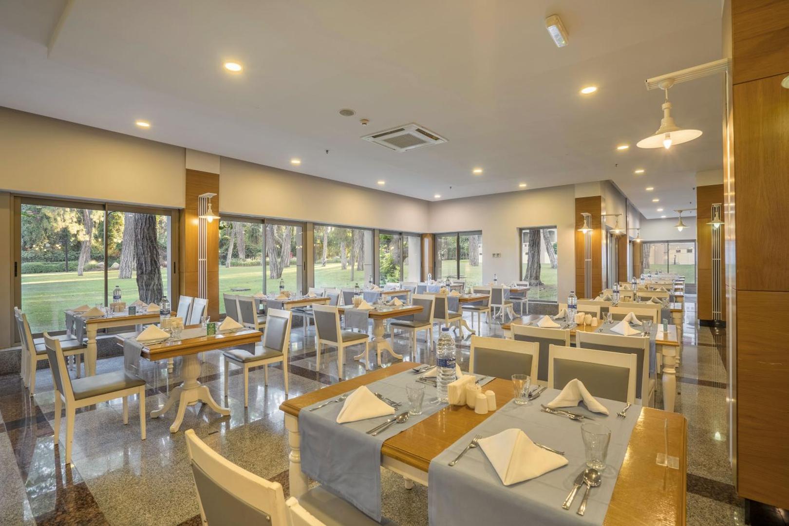 Mirada Del Mar Hotel gaiša restorāna zāle ar lieliem logiem uz dārzu un servētiem galdiem Kemeras piekrastē.