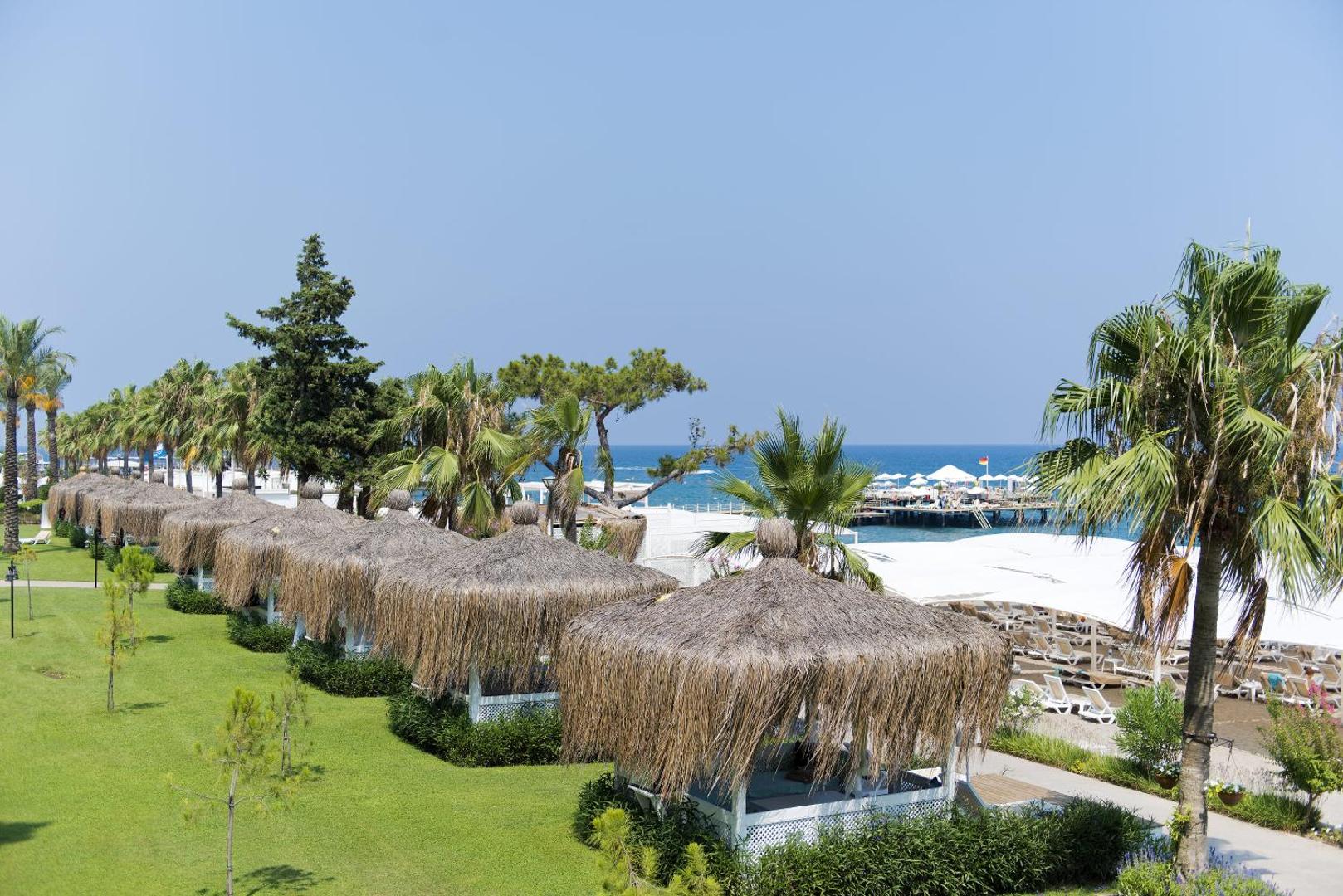 Mirada Del Mar Hotel pludmales atpūtas zonas ar salmu jumtiem un palmām — skats uz jūru un piestātni Kemeras kūrortā