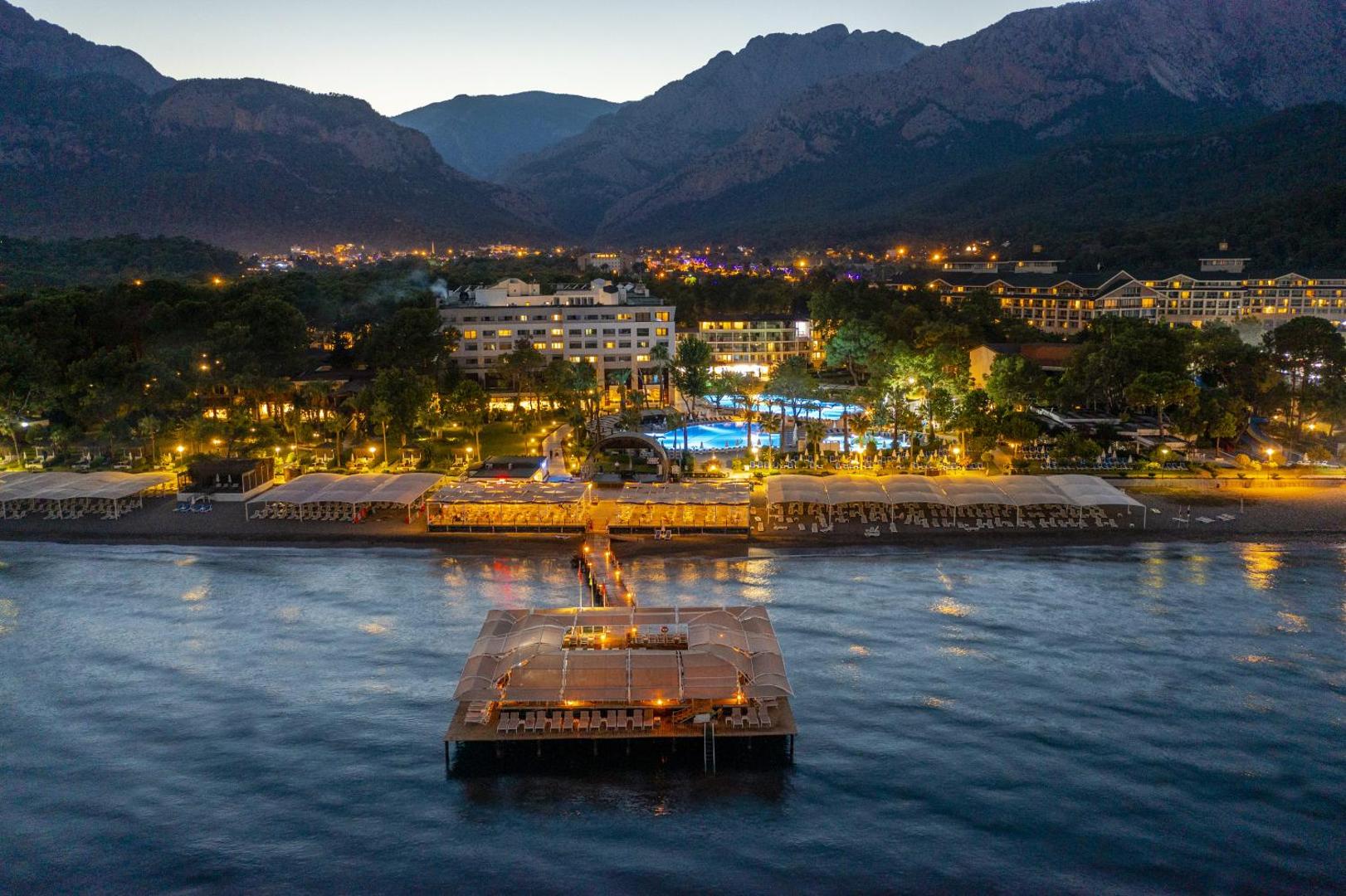 Mirada Del Mar Hotel vakara apgaismojums Kemeras piekrastē — izgaismota piestātne, baseini un viesnīcas korpuss uz kalnu fona