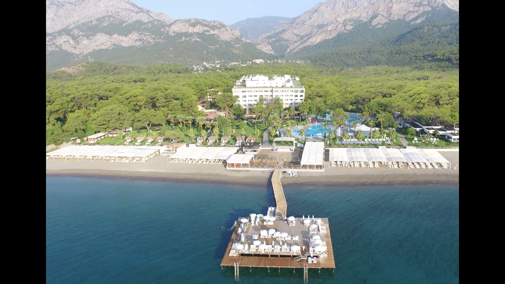 Mirada Del Mar Hotel panorāmas skats uz pludmali un piestātni ar saulessargiem — fonā redzami augsti kalni un mežs