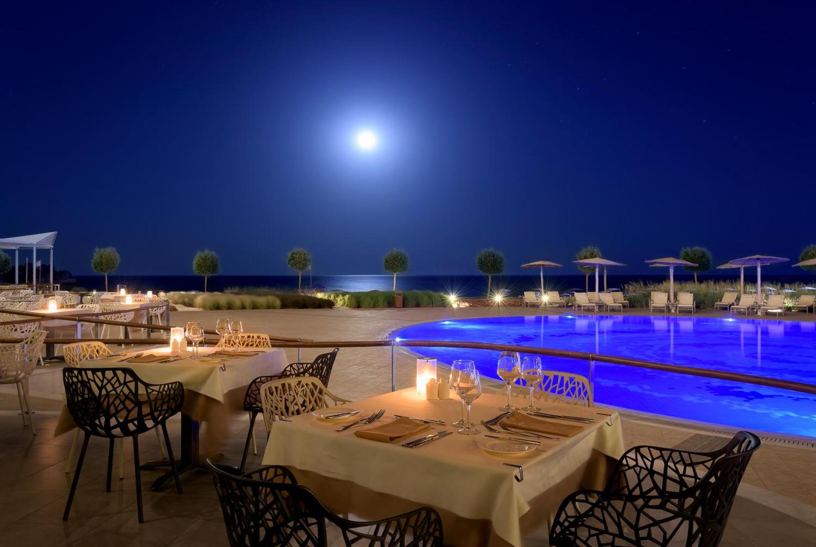 Elysium Resort & Spa āra restorāns un izgaismots baseins naktī zem pilnmēness pie jūras krasta Rodā