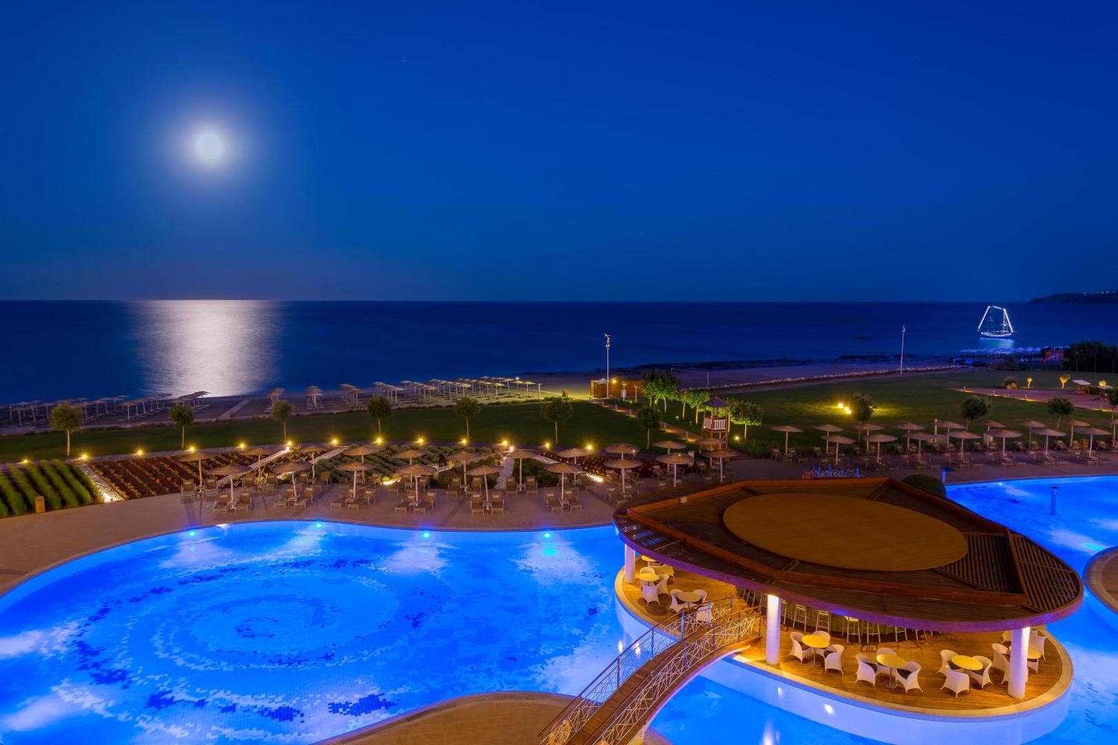 Elysium Resort & Spa baseins un bārs naktī ar izgaismotu tiltiņu un pilnmēnesi virs Vidusjūras Rodā