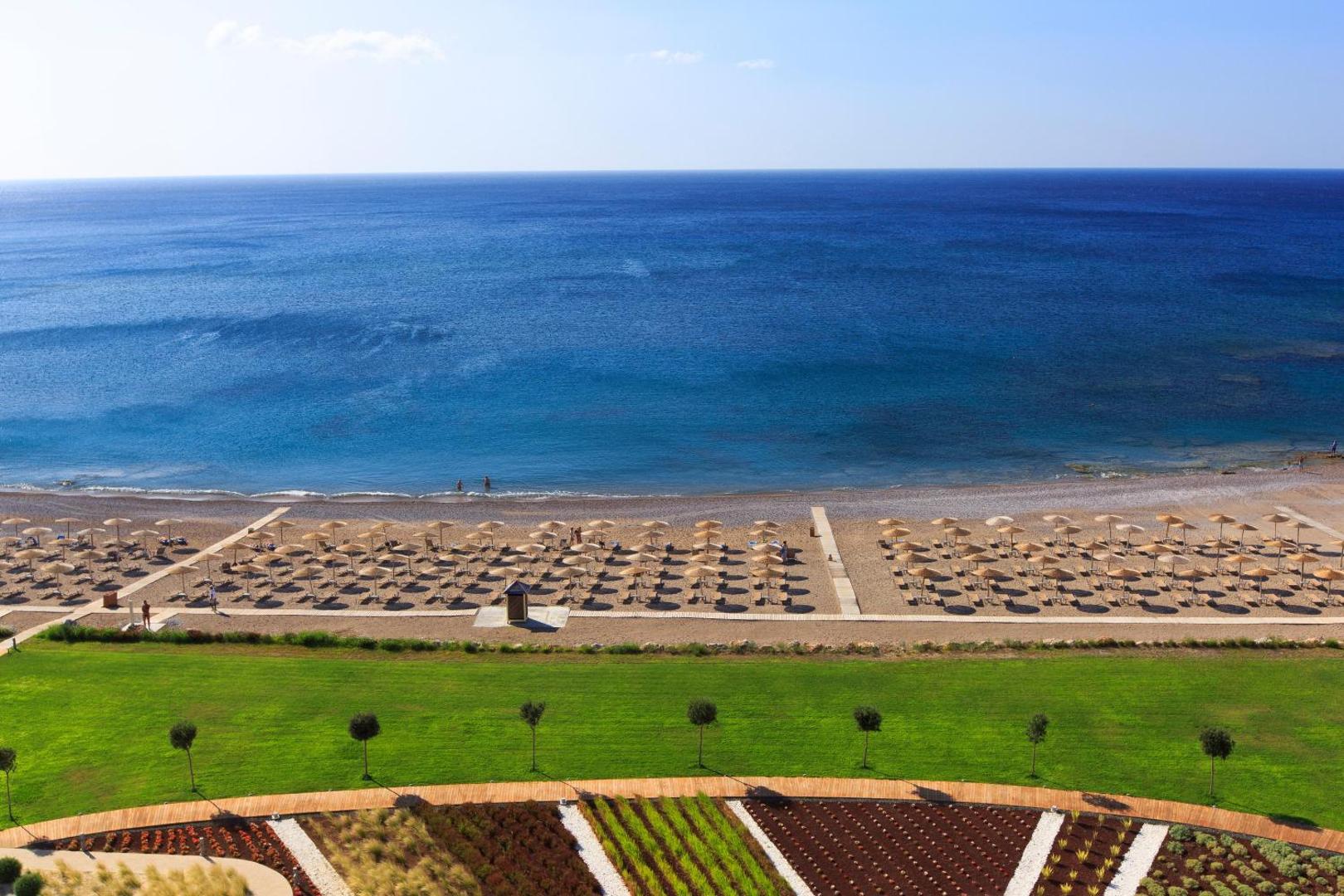 Elysium Resort & Spa plašā privātā pludmale Rodas salā ar saulessargiem, zālāju un skatu uz zilo Vidusjūru no augšas