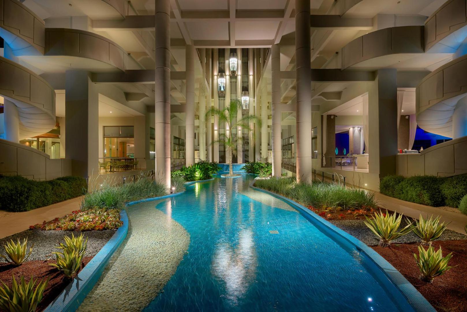 Elysium Resort & Spa iekšpagalma dekoratīvais baseins ar palmu un modernām kolonnām — vakarā izgaismota atpūtas zona Rodas kūrortā