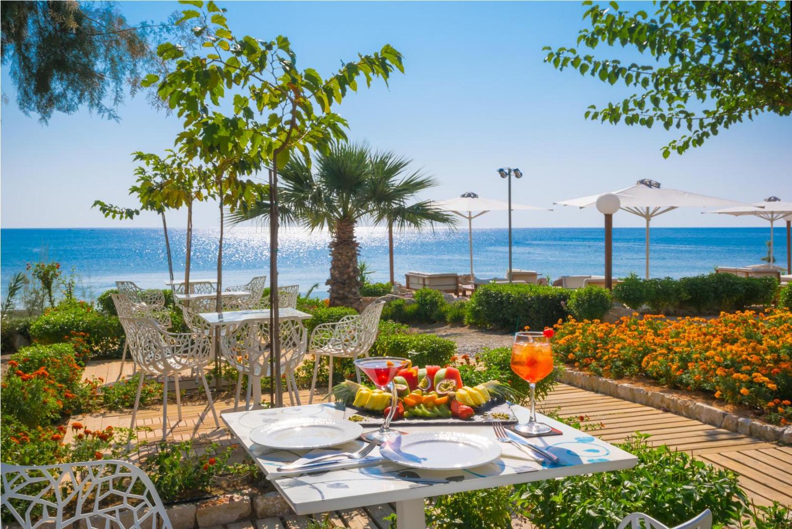 Elysium Resort & Spa dārza terase ar augļiem, kokteiļiem un baltiem galdiņiem ziedošu puķu un palmu ielenkumā pie jūras