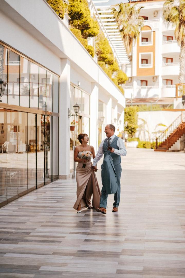 Laimīgs pāris svinīgā apģērbā pastaigājas pa Castival Hotel viesnīcas teritoriju — gaiša terase un moderna arhitektūra fonā