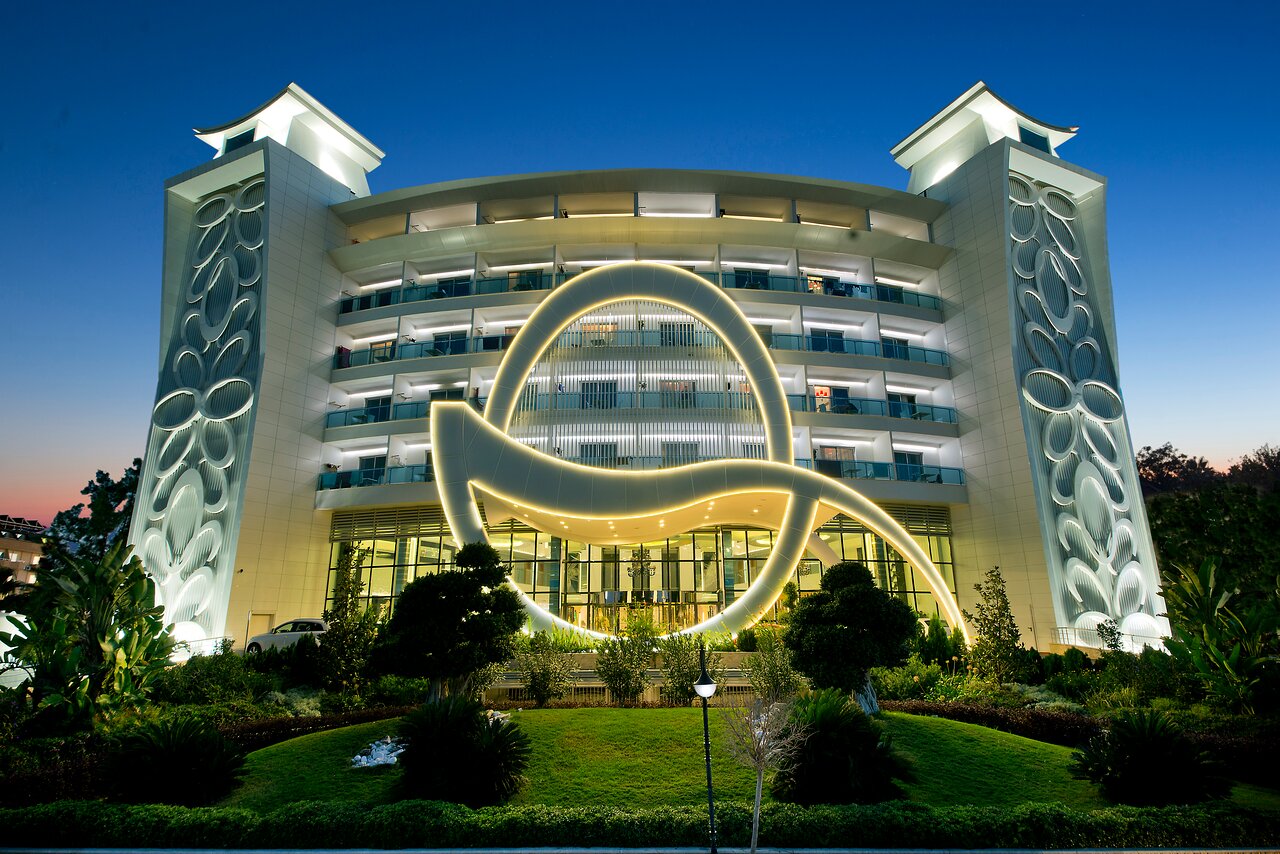 Q Premium Resort Hotel galvenā ēka vakarā ar izgaismotu modernu fasādi un ieeju Alānijā, Turcijā — arhitektūras detaļas un dārzs