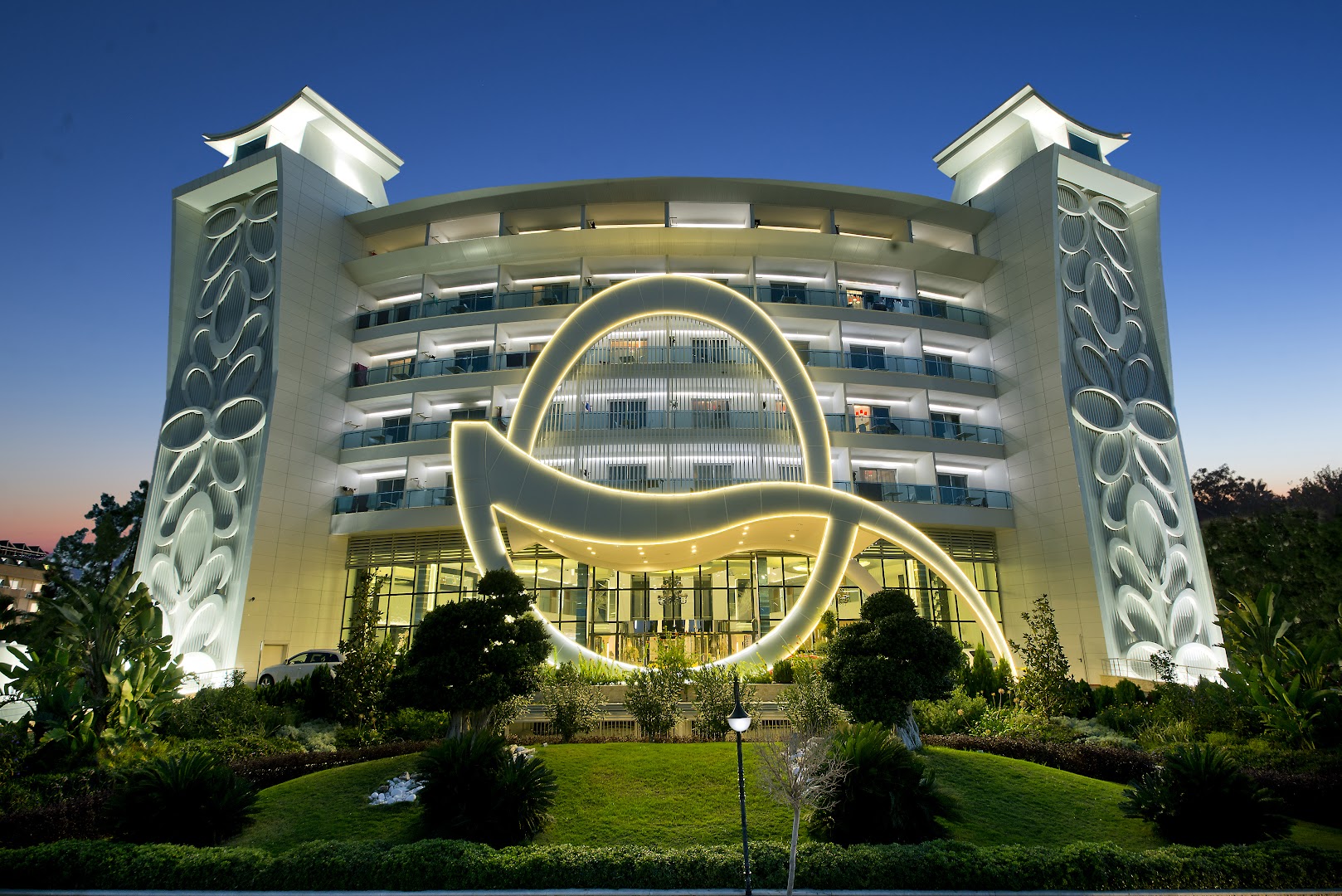 Q Premium Resort Hotel fasādes tuvplāns krēslas stundā ar dekoratīvu apgaismojumu un koptu zālienu priekšplānā