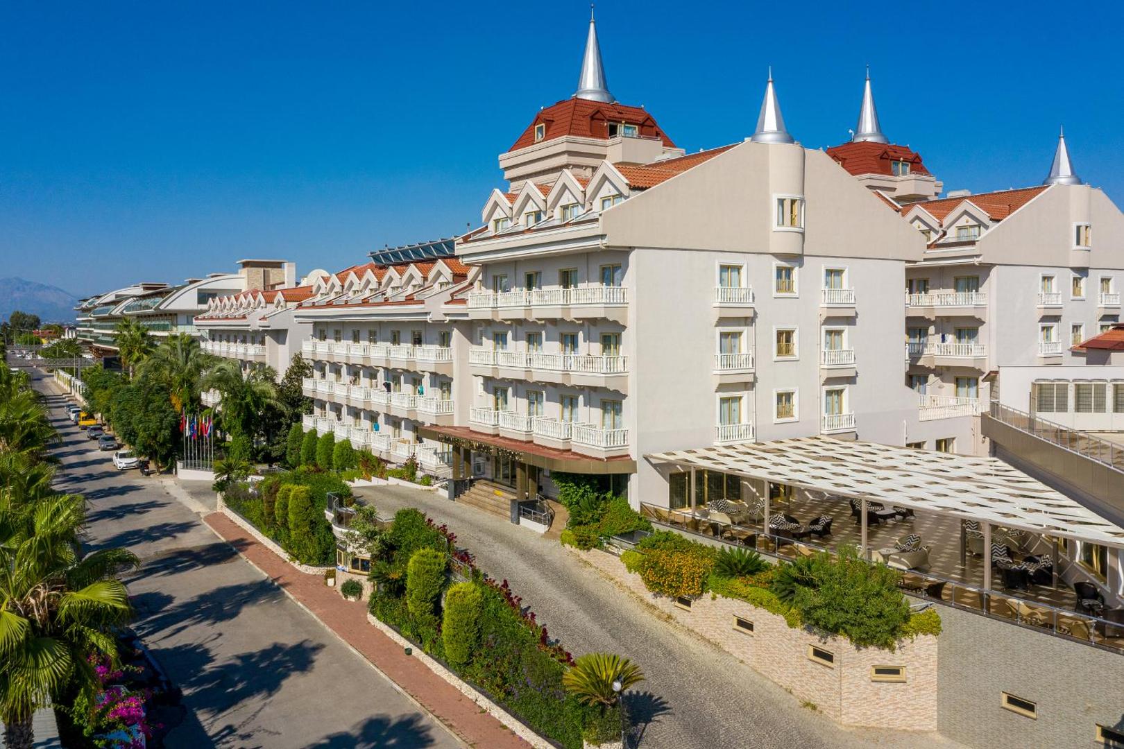 Aydinbey Famous Resort galvenā ēka ar raksturīgajiem smailajiem torņiem un balkoniem saulainā dienā — ieeja un āra terase