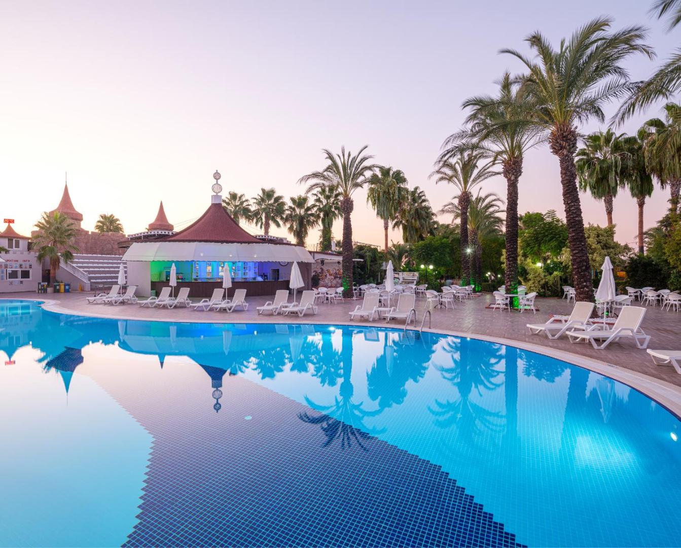 Aydinbey Famous Resort baseina zona ar bāru un atpūtas krēsliem saulrietā — palmu atspulgi dzidrajā ūdenī
