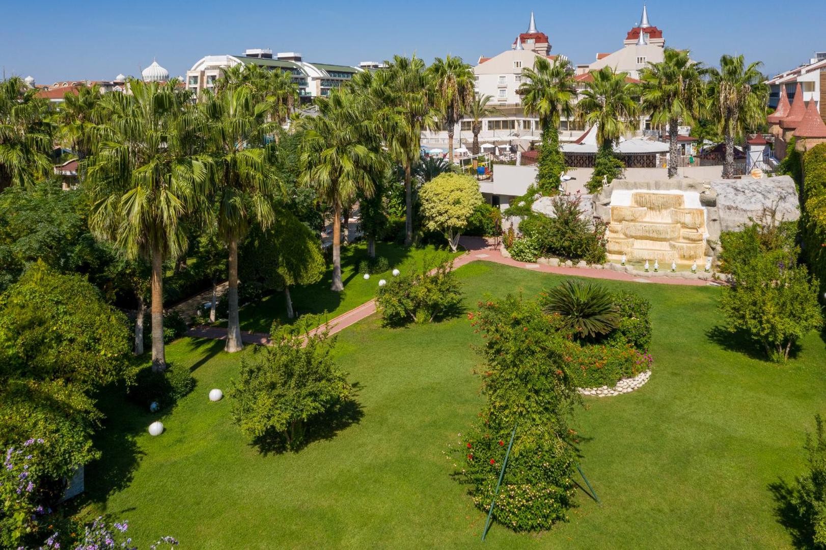 Aydinbey Famous Resort zaļais dārzs ar koptu zālienu, palmām un dekoratīvu ūdenskritumu — mierīga atpūtas zona viesnīcas teritorijā