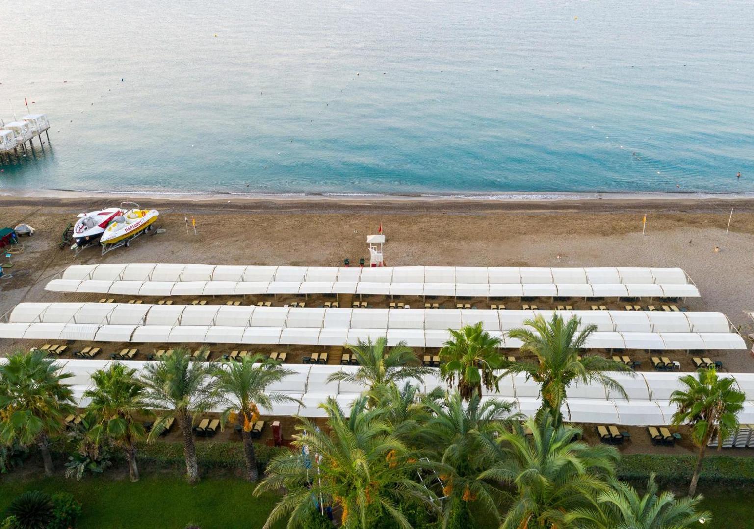 Aydinbey Famous Resort pludmales zona ar palmām un nojumēm — skats uz Vidusjūru un smilšu joslu pie viesnīcas