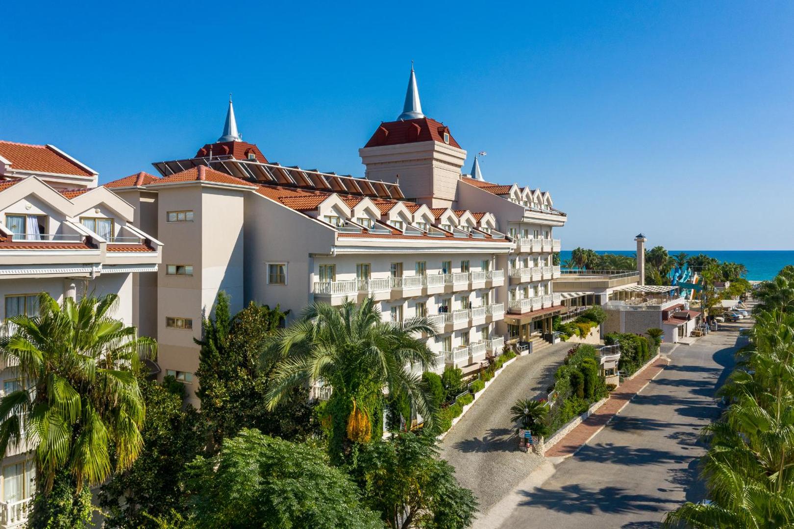 Aydinbey Famous Resort viesnīcas korpuss ar skatu uz Vidusjūru — palmas, sakopta teritorija un saulains debess jums