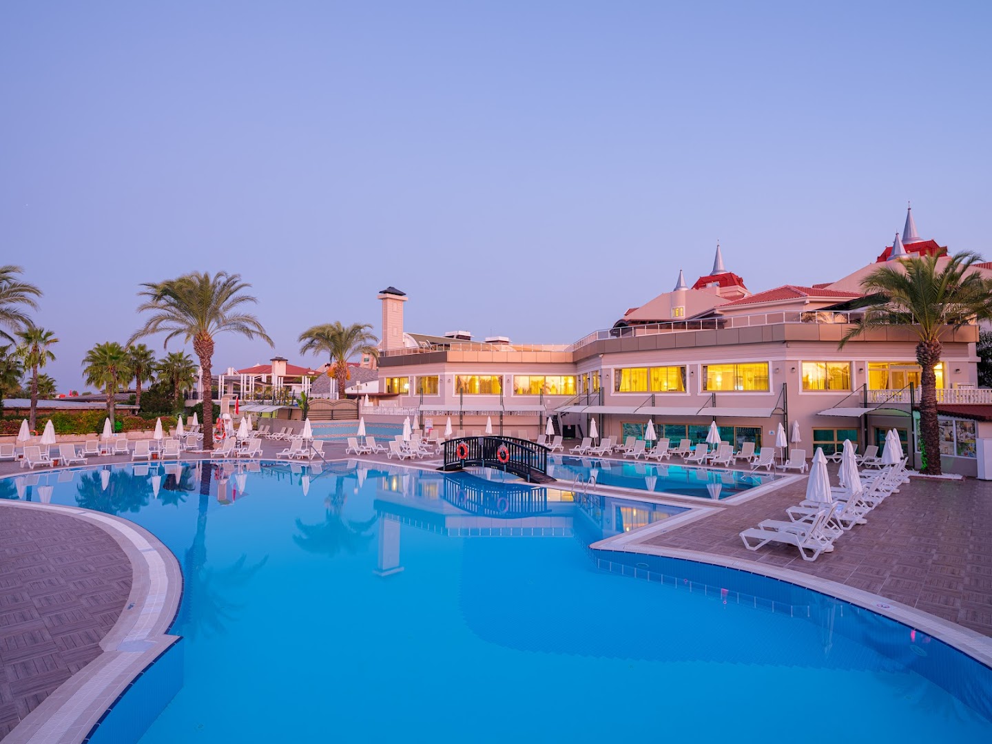 Aydinbey Famous Resort galvenais baseins un viesnīcas ēka krēslas stundā ar izgaismotiem logiem un tiltiņu Belekā