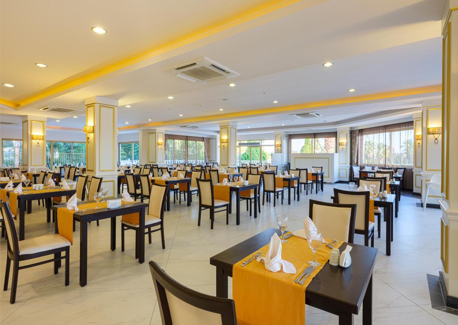 Aydinbey Famous Resort galvenais restorāns ar gaišu interjeru un dzelteniem akcentiem — plaša ēdamzāle ar servētiem galdiem