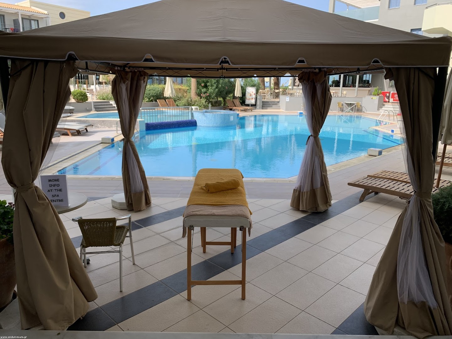 Pearl Beach āra baseins un masāžas telts dārzā — skats no spa zonas uz saulaino terasi un zilo ūdeni Krētā