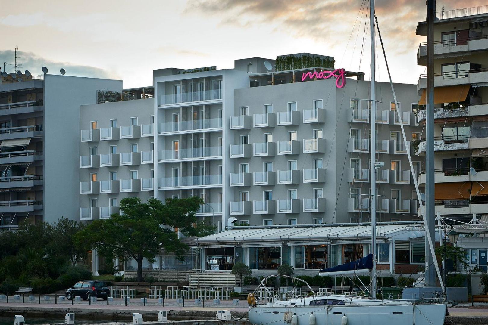 Moxy Patra Marina viesnīcas skats dienas gaismā no krastmalas — moderna daudzstāvu ēka ar balkoniem un jahtu piestātni priekšplānā