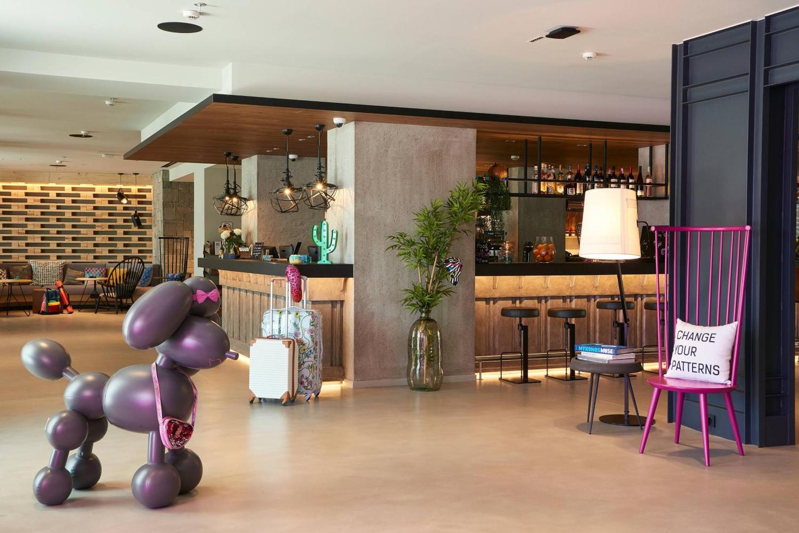 Plašs Moxy Patra Marina vestibils ar dizaina mēbelēm, košu rozā krēslu un māksliniecisku pūdeļa skulptūru priekšplānā