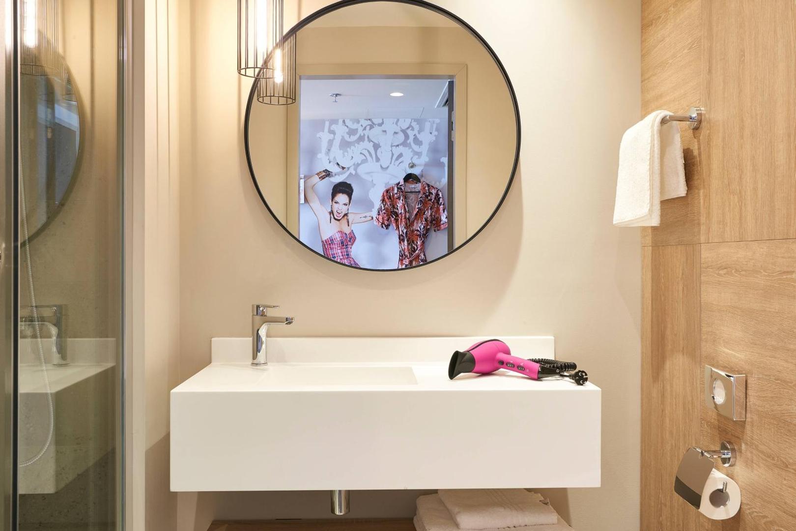 Moxy Patra Marina numura vannasistaba ar modernu izlietni, apaļu spoguli un koši rozā fēnu, atspoguļojot viesnīcas rotaļīgo dizainu.