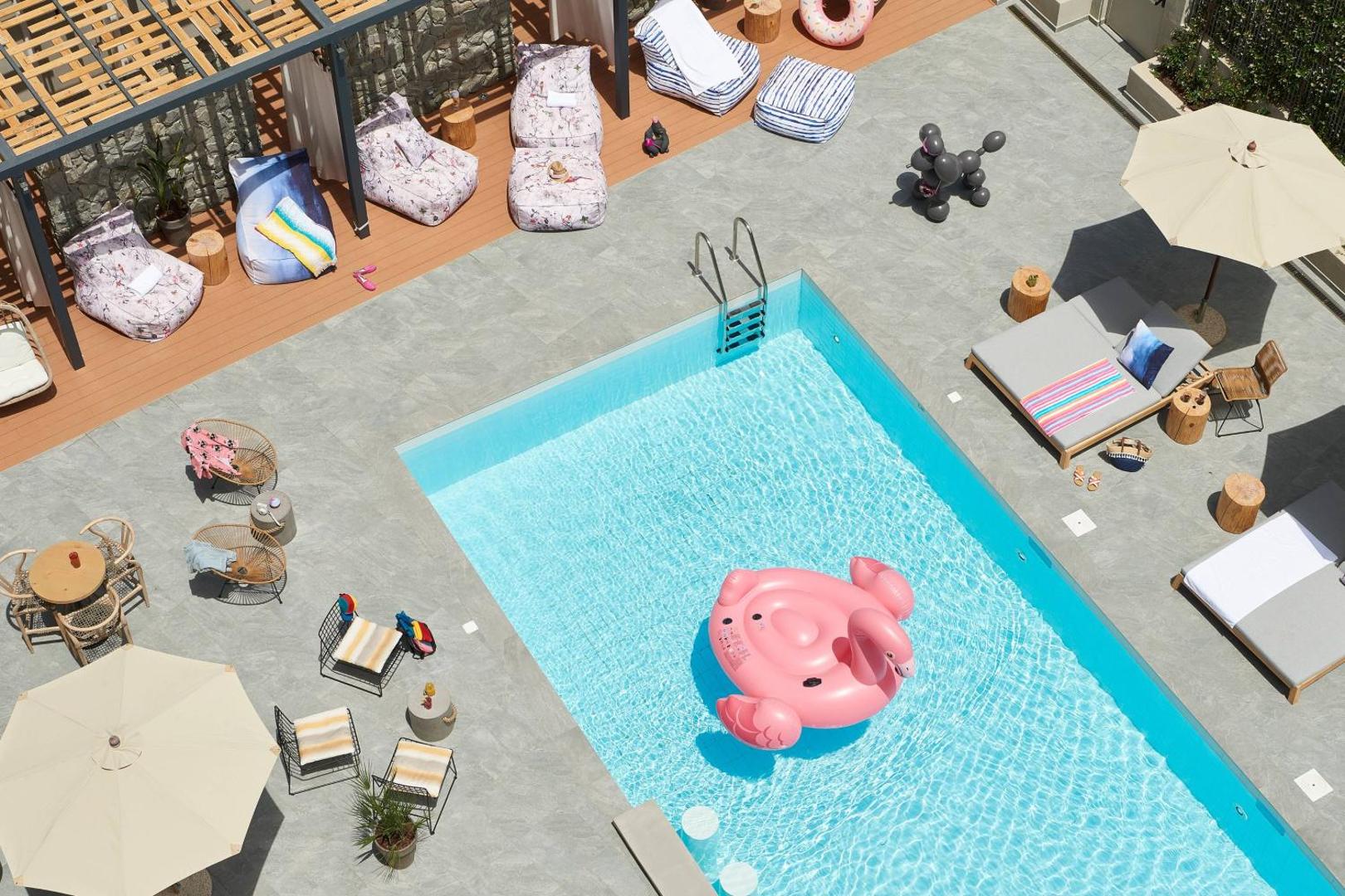 Moxy Patra Marina viesnīcas āra baseins no augšas Patrā, Grieķijā — atpūtas zona ar krāsainiem sēžammaisiem, saulessargiem un piepūšamo rozā flamingo