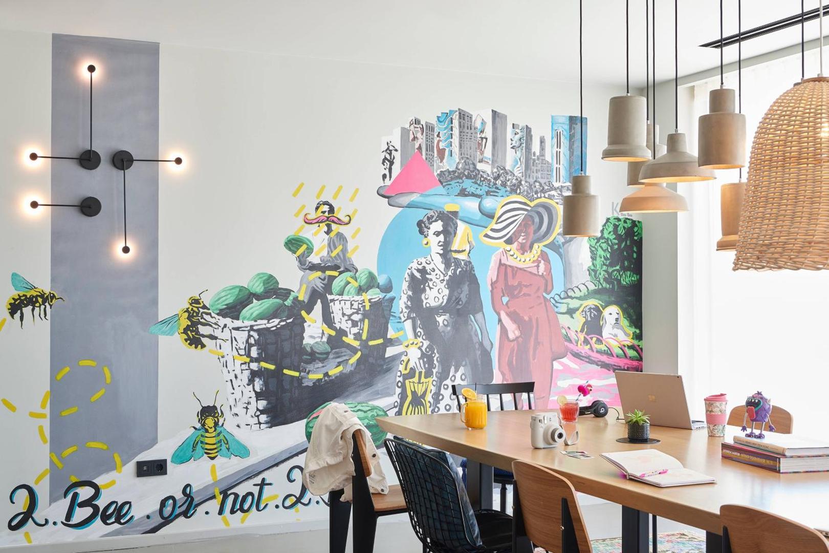 Moxy Patra Marina kopstrādes zona ar lielu koka galdu, modernu sienas gleznojumu un dizaina lampām radošai darba videi.