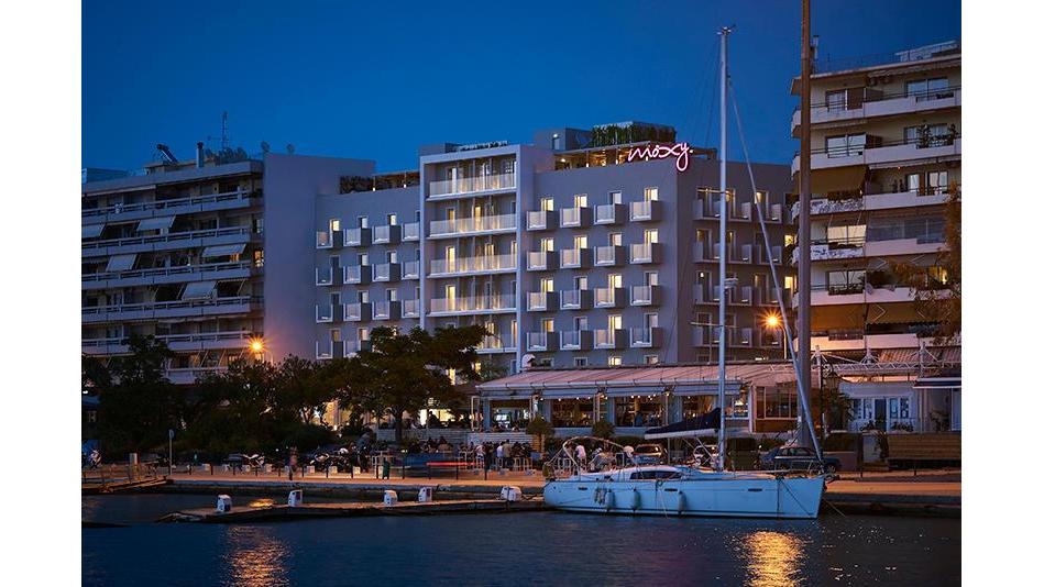 Moxy Patra Marina ēka krēslas stundā no ūdens puses — redzami numuri ar balkoniem un piekrastes restorāns blakus jahtām