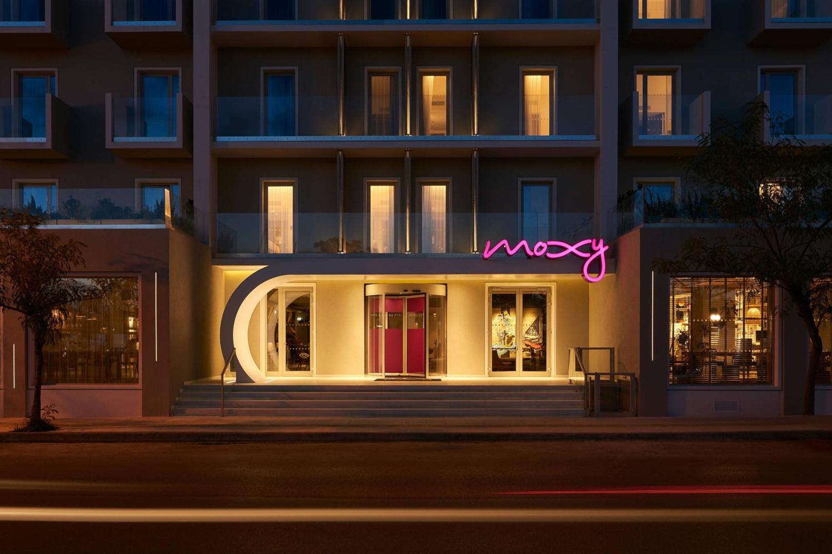 Moxy Patra Marina galvenā ieeja naktī ar rozā neona logotipu un modernu arhitektūru — apgaismotas kāpnes un stikla durvis