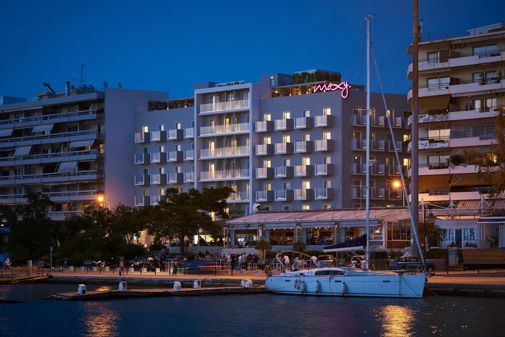 Moxy Patra Marina viesnīcas fasāde vakarā pie jahtu piestātnes Patrā, Grieķijā — pilsētas ainava ar izgaismotiem numuru balkoniem un jahtu priekšplānā