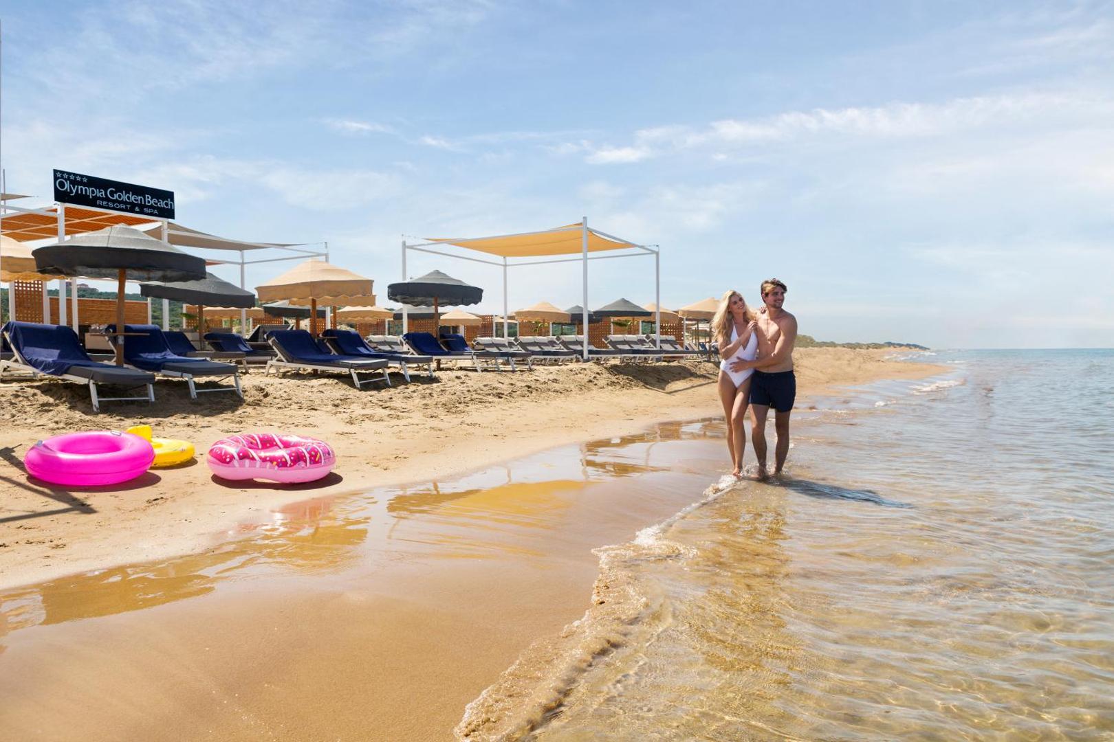 Olympia Golden Beach Resort & Spa privātā pludmale Peloponēsā ar pāri, saulessargiem, sauļošanās krēsliem un rozā piepūšamajiem riņķiem smiltīs.