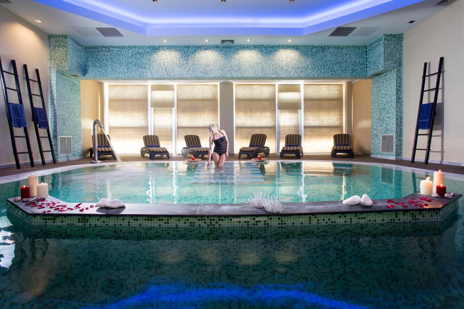 Olympia Golden Beach Resort & Spa iekštelpu baseins spa centrā ar atpūtas krēsliem un dekoratīvām svecēm — relaksācijas zona viesiem