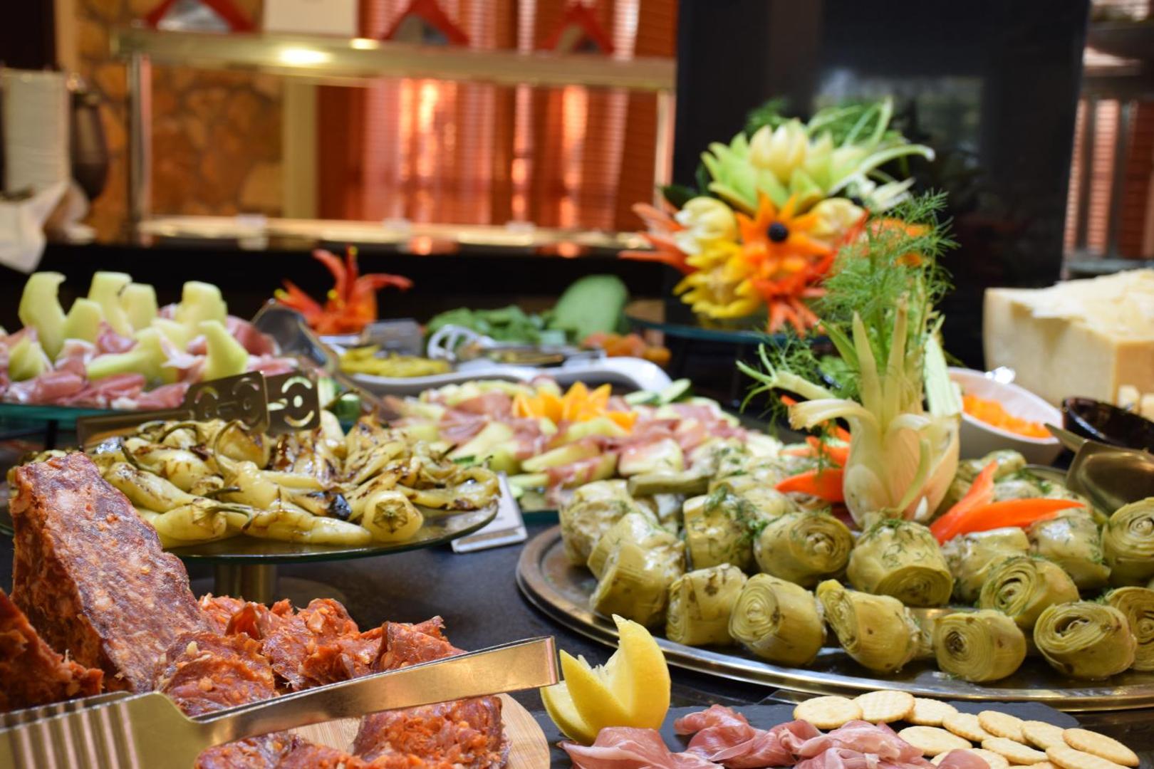 Olympia Golden Beach Resort & Spa restorāna bufetes galds ar daudzveidīgām uzkodām, artišokiem, gaļas izstrādājumiem un dekoratīviem dārzeņu griezumiem.