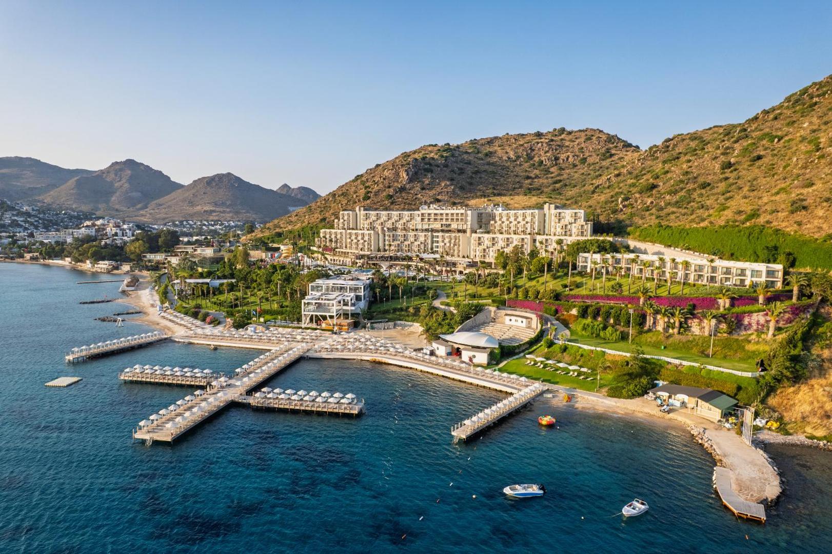 Kefaluka Resort Hotel skats no putna lidojuma uz pludmali un piestātnēm Bodrumā, Turcijā — kalnu ieskauta piekrastes viesnīca ar koptu dārzu