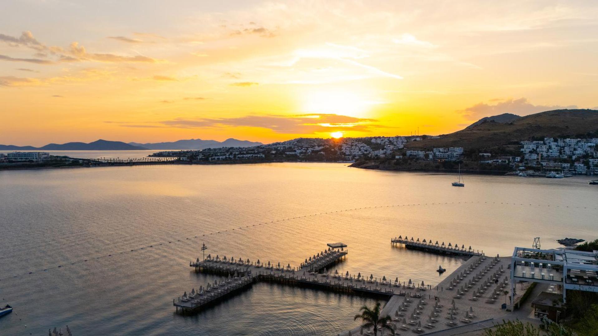 Kefaluka Resort saulrieta skats uz Egejas jūras līci un koka piestātni Bodrumā — zeltītas debesis virs kalniem un piekrastes pilsētas