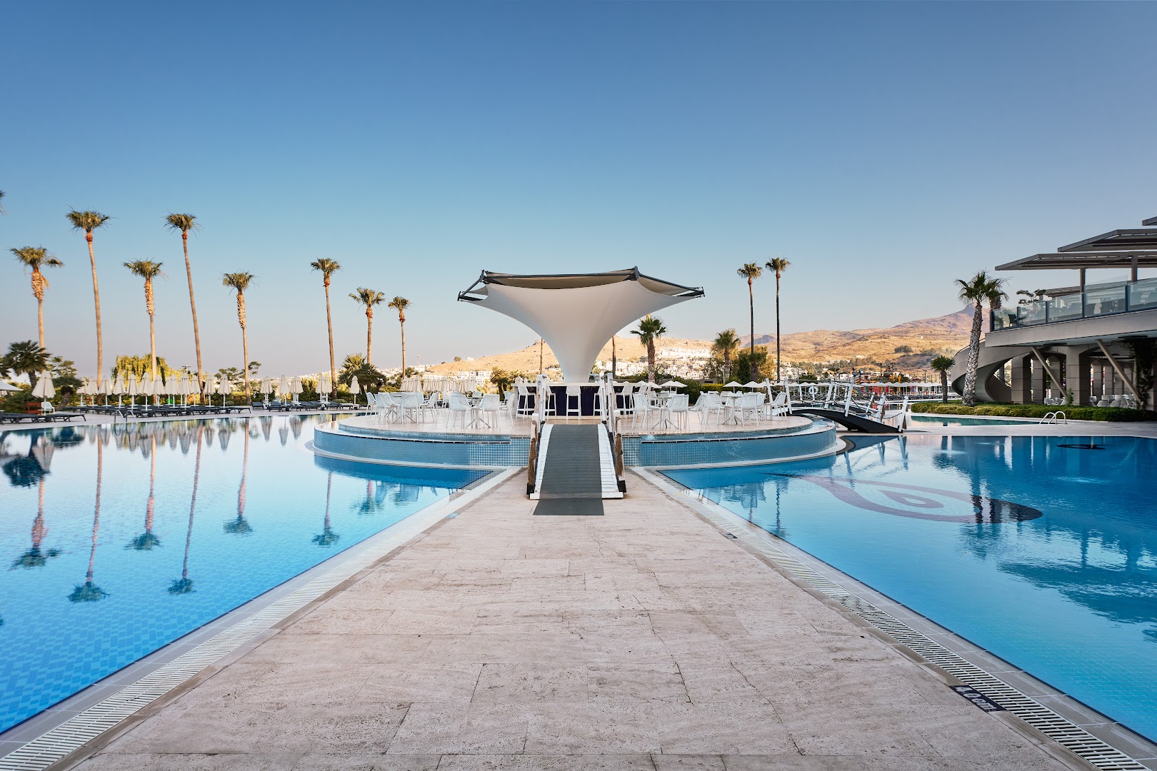 Kefaluka Resort Hotel baseina bārs ar modernu tentu un tiltiņu — skats uz kalniem un dzidri zilo ūdeni