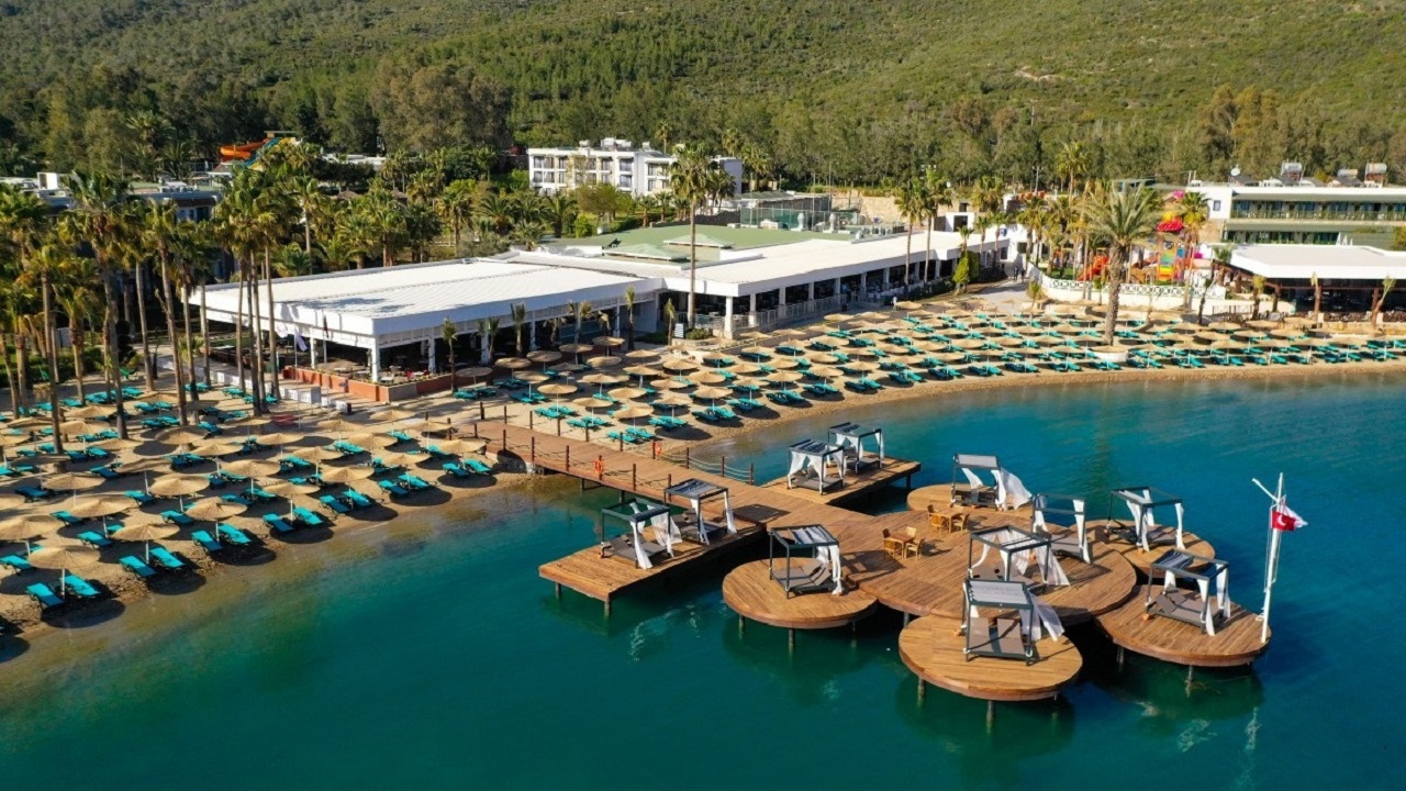 Crystal Green Bay Resort panorāmas skats uz privāto pludmali, koka laipu un viesnīcas ēkām priežu meža ieskautā līcī Bodrumā