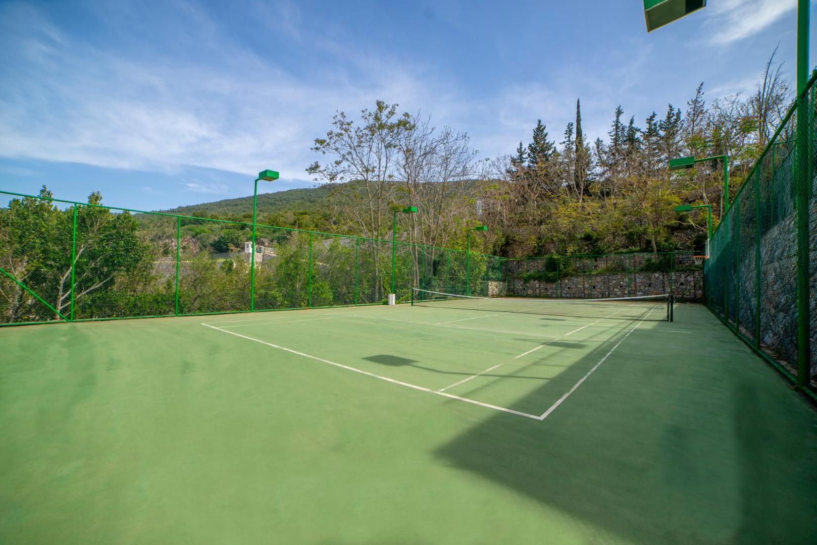 Duja Bodrum tenisa korts ar cieto segumu, ko ieskauj zaļš žogs un kalnains apvidus ar priedēm Turcijā