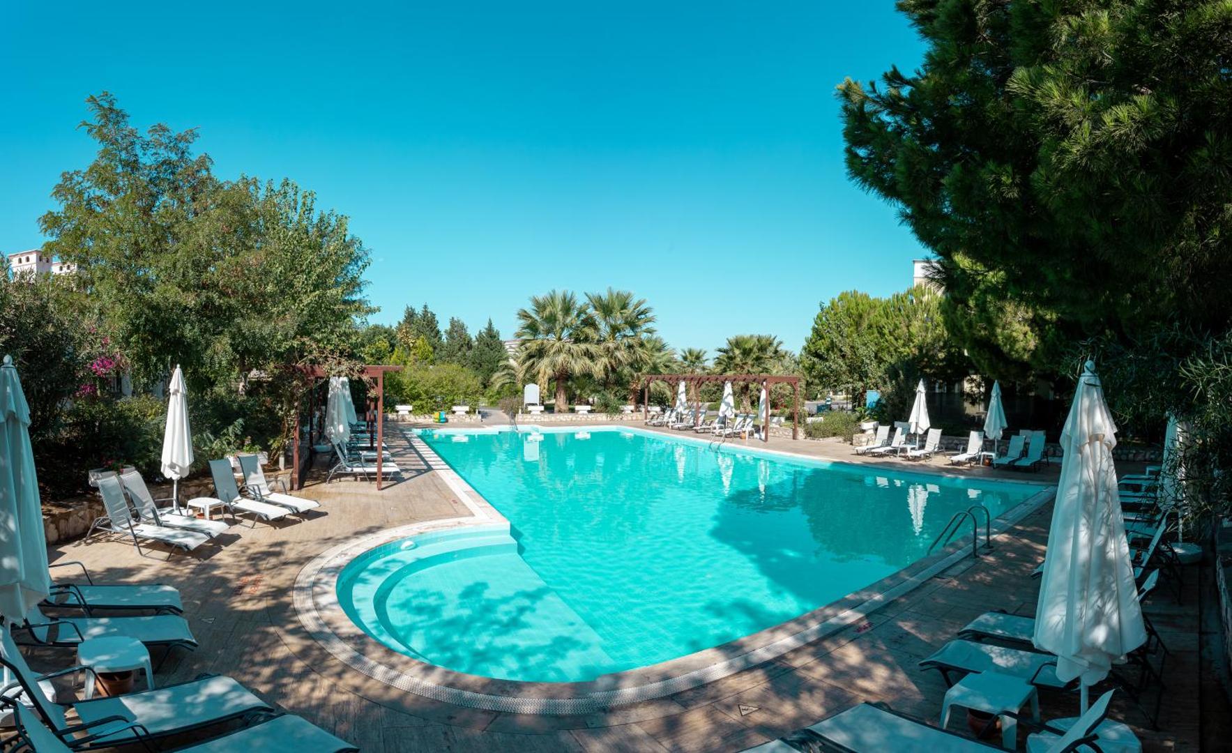 Anadolu Hotels Didim Club āra baseins ar dzidru ūdeni, ko ieskauj koki, palmas un balti saulessargi ar ērtiem atpūtas krēsliem.