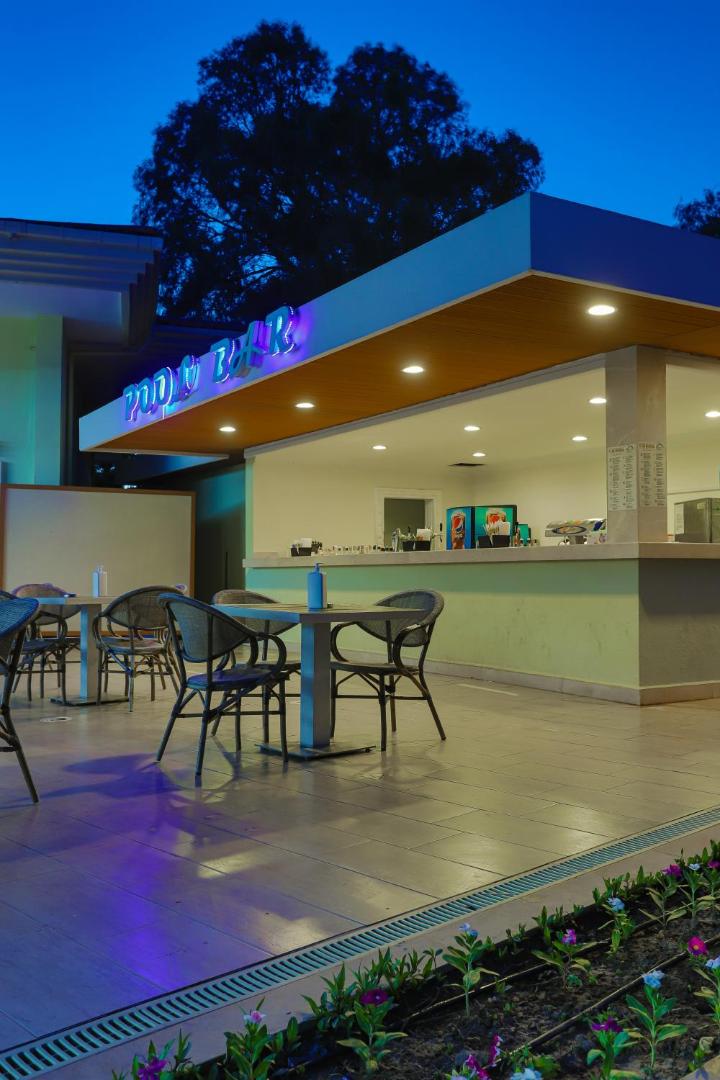 Anadolu Hotels Didim Club baseina bārs vakarā ar apgaismojumu, āra galdiņiem un krēsliem mājīgā atmosfērā