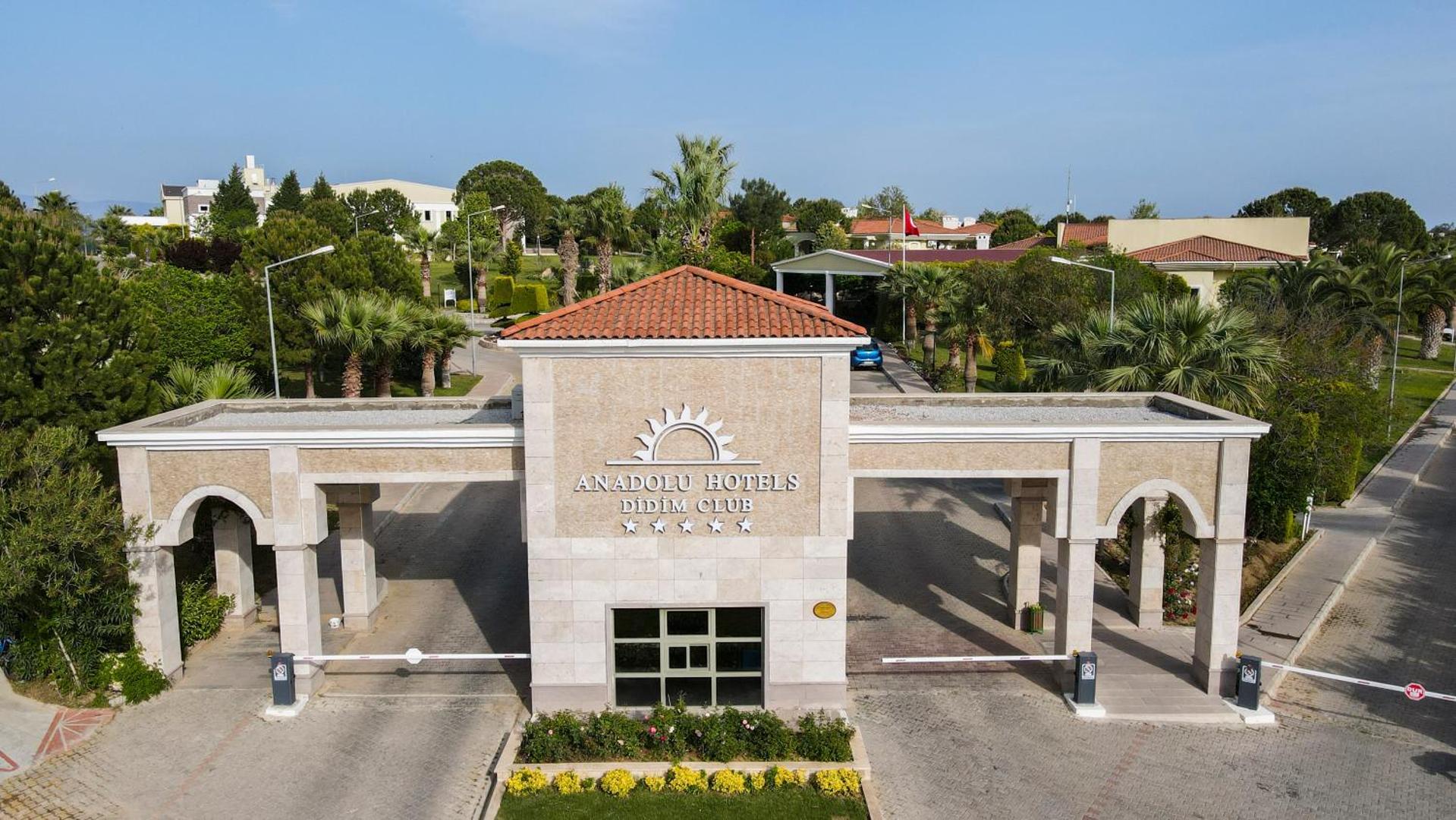 Anadolu Hotels Didim Club galvenā iebrauktuve ar viesnīcas nosaukumu un piecu zvaigžņu simboliku, ko ieskauj zaļi koki un palmas