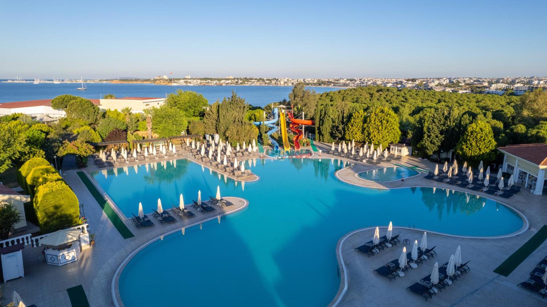 Anadolu Hotels Didim Club baseina zona saulrietā ar skatu uz Egejas jūru un pilsētas panorāmu, apkārt zaļojoši koki un atpūtas krēsli.
