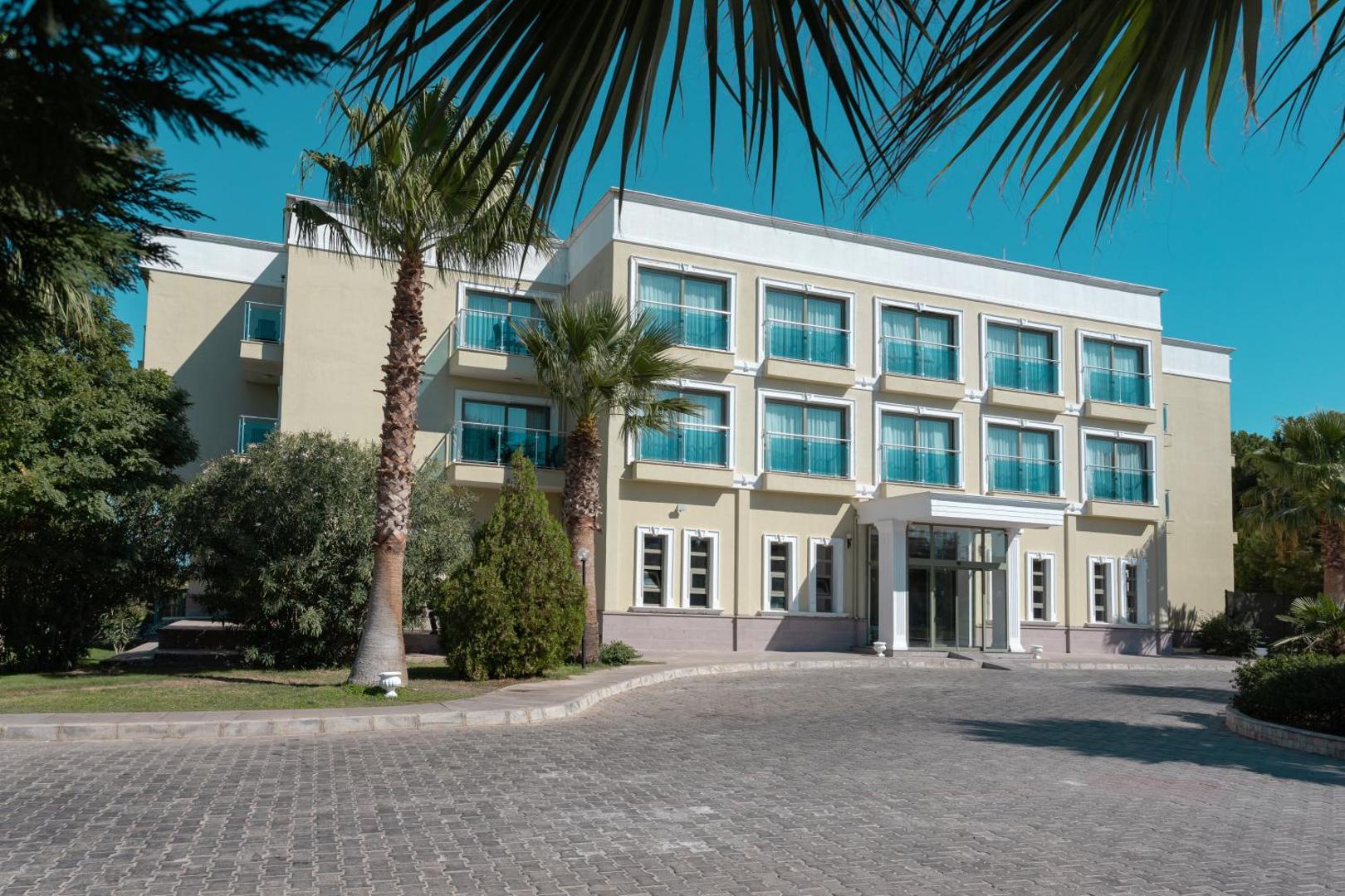Anadolu Hotels Didim Club galvenā korpusa ēka ar balkoniem, palmām un bruģētu piebraucamo ceļu skaidrās debesīs