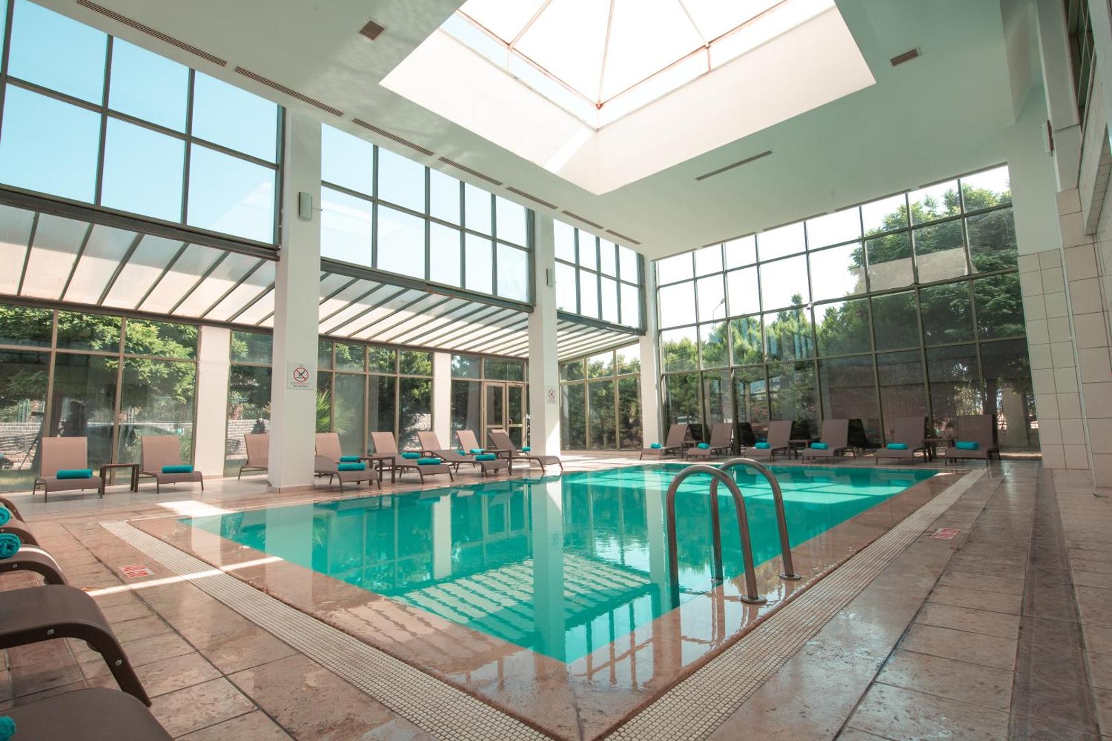 Anadolu Hotels Didim Club iekštelpu baseins ar stikla sienām un jumta logu, apkārt izvietoti atpūtas krēsli ar tirkīza krāsas dvieļiem.
