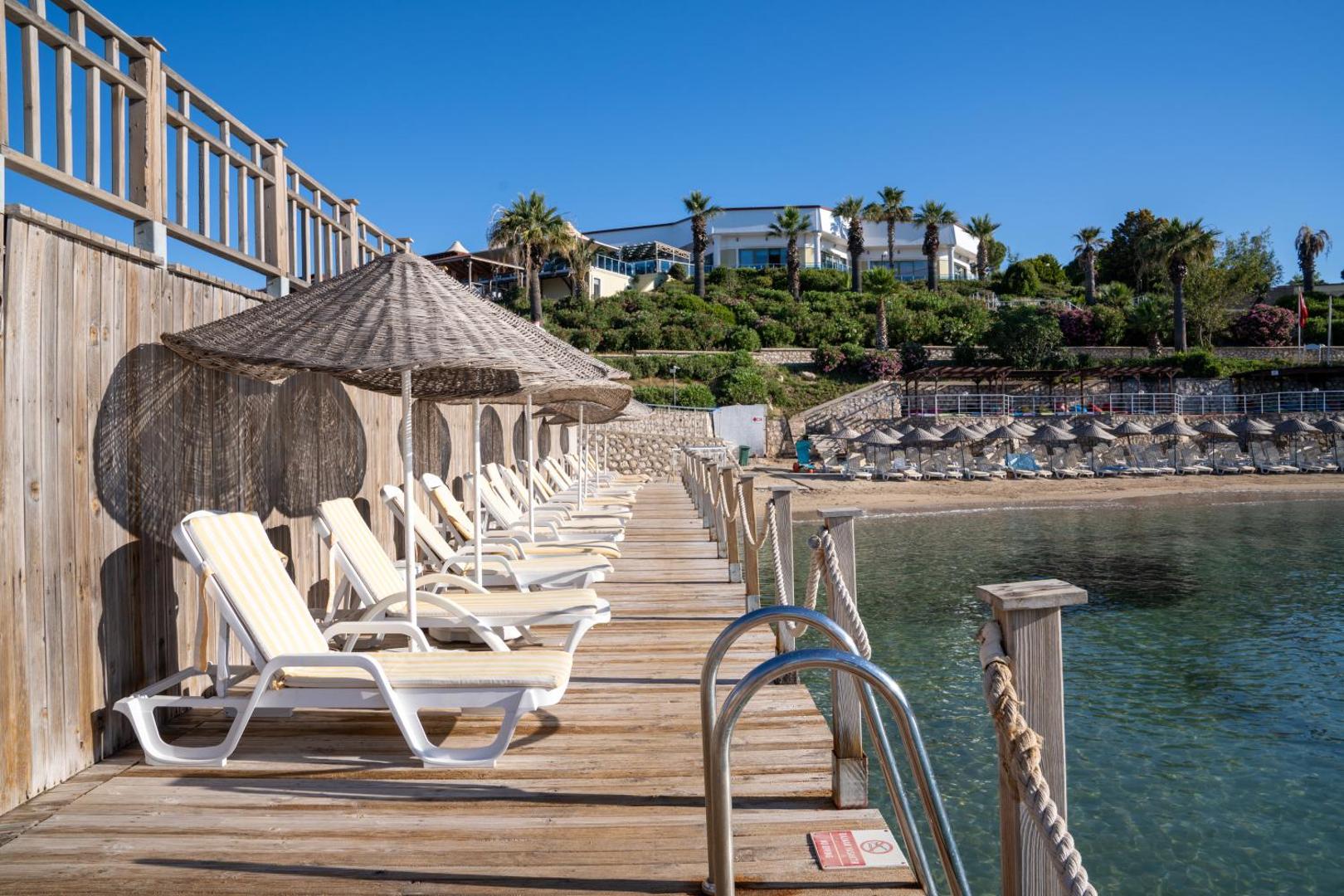 Anadolu Hotels Didim Club koka laipa ar sauļošanās krēsliem un saulessargiem pie jūras, skats uz viesnīcas galveno ēku kalnā